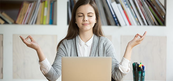 Person sitting in front of a laptop with bookshelves in the background. The person's hands are raised with thumb and forefingers touching and their eyes are closed.
