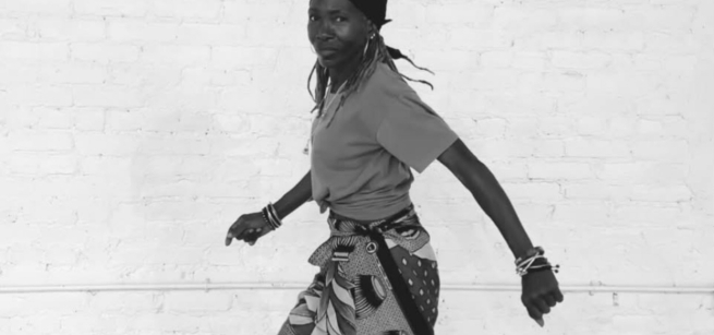 black and white photo of a Black woman standing in a dance pose wearing a boldly printed cloth skirt with a solid t-shirt