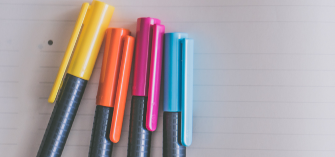 Four colorful markers, yellow, orange, pink, and teal, lined up on a sheet of lined paper.