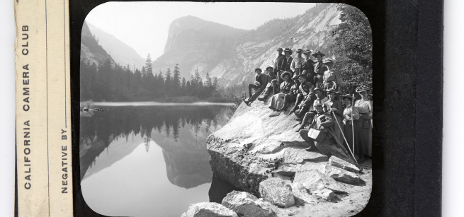 A black and white slide from the California Camera Club, a group of men and women are gathered on the shore of a lake, they are wearing clothing typical of the early 1900s
