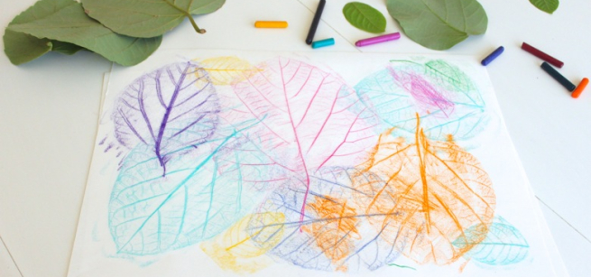 Green leaves are on the table beside a white paper with colorful leaf rubbings featured on the top of the page. 