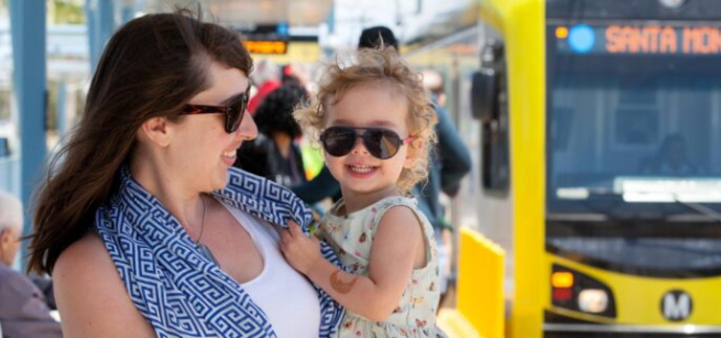 An adult wearing sunglasses holds a child wearing sunglasses in their arms in front of the Santa Monica Metro bus.