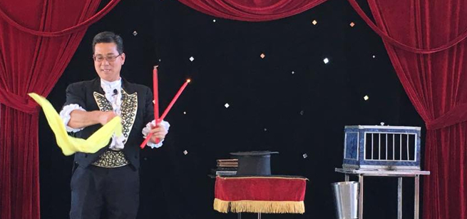 Magician waving a scarf and wand, near a table with a top hat and rabbit cage.