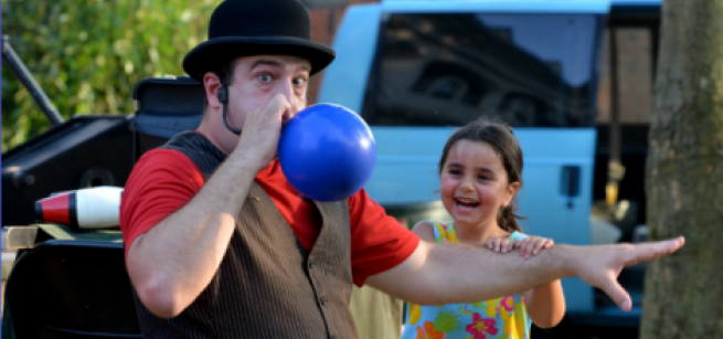 Magician Amazing Dave blowing up a balloon as a smiling child looks on.