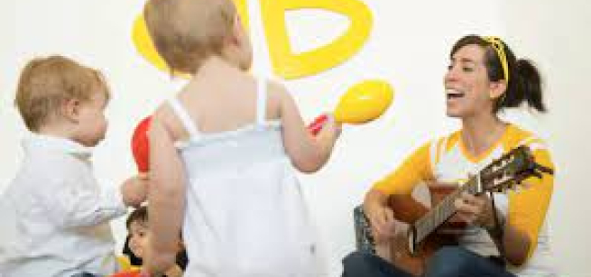 An adult female sitting on the floor playing a guitar and two toddlers watching her,