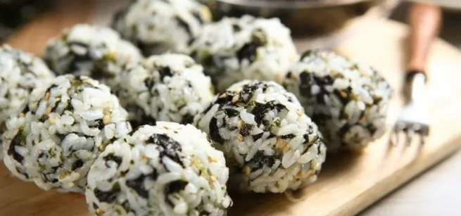 A group of rice balls mixed with seaweed and sesame seeds are arranged on a wooden board with a fork in the background.
