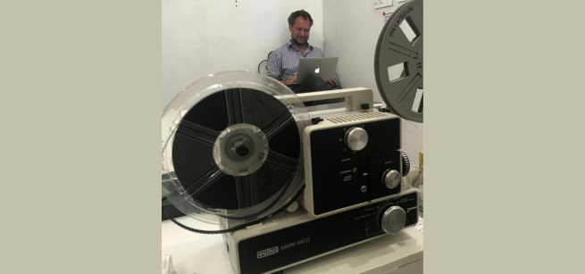 8mm film projector in the foreground with a film instructor working on a laptop in the background. 