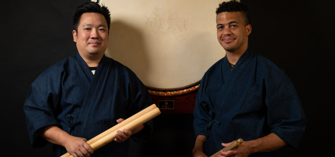 Two BIPOC men in blue wrap coats, holding drumsticks, standing in front of a large taiko drumhead