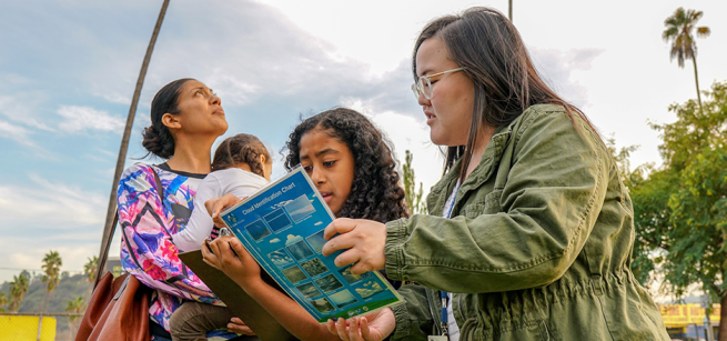 In an outdoor area with palm trees, a female adult with straight dark hair, eyeglasses and a green jacket is holding an instruction guide on clouds with a young girl with curly brown hair. Another female with lght brown curly hair in bun and holding a toddler over her shoulder is to the left of them looking at a blue sky with partial cloud cover and the sun above.