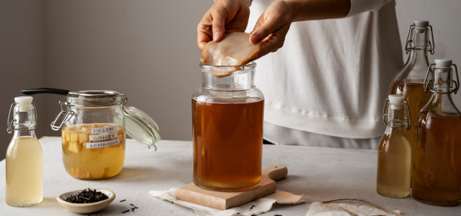 Side view of person in a white sweatshirt holding a round scoby over a big jar on a white table with several various-sized glass containers with yellow and brown colored tea. Also shows a white small shallow bowl next to a wooden spoon with black looseleaf tea.