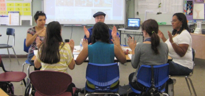 A group of adults sitting in a circle in chairs, drumming a rhythm with their hands