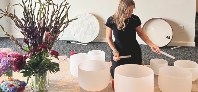 Blonde woman in all black sitting in the middle of six variously sized glass bowls, looking down with the playing sticks and a large lavender bouquet in a glass to the left of the image.   