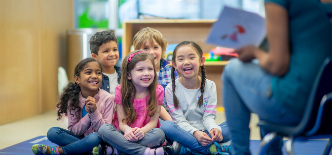 Children sit on floor and look up to adult who is reading. 