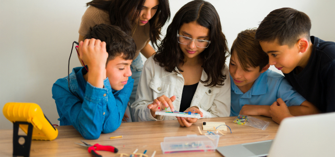 Five teens are gathered around a circuit board and looking at it with interest. They are surrounded by plyers and other tools.