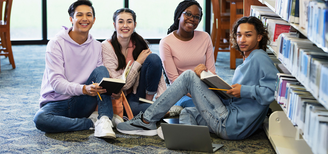 A diverse group of teens sits together in the library, smiling