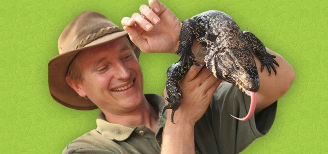 A smiling man named Thor wearing a safari hat looks at a huge lizard laying on his arm.