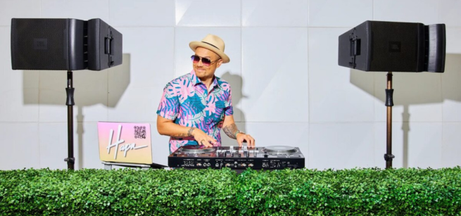 DJ in a Hawaiian shirt and hat stands behind a DJ setup