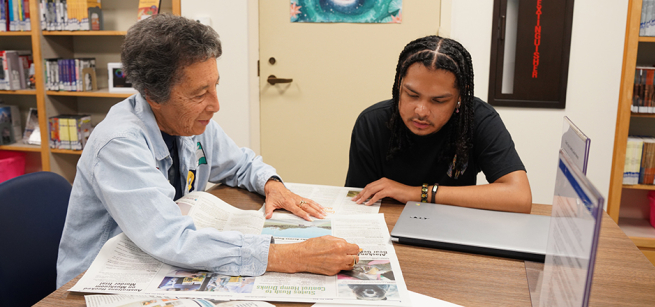 A person helping another person read a newspaper.