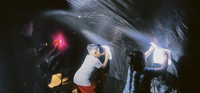 Two kids holding paper up to the sides of the black plastic wall of the planetarium while an adult shines a flashlight so they can see