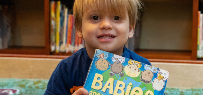 Toddler seated on a colorful rug in children's section of library, holding a book with illustrations of babies, titled "Babies."