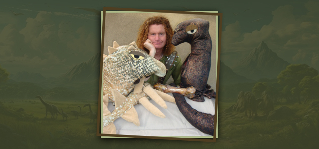 Man with red hair sitting between two dinosaur puppets. 