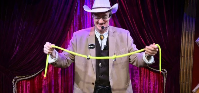 Smiling man with a cowboy hat looks down at a rope in between his hands ready for the next magic trick. 