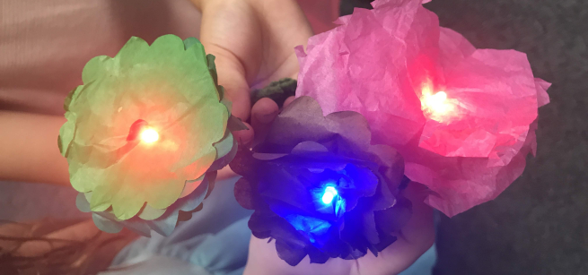 Close-up of children's hands showing the camera their lit-up tissue paper flowers in colors green, blue and pink.