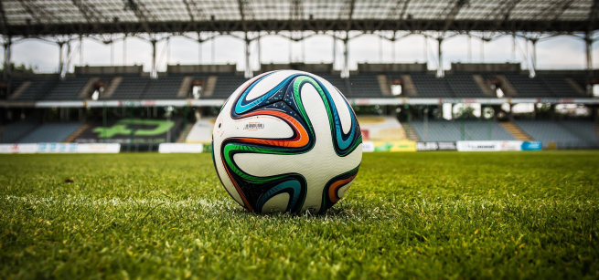 an orange, blue, green, and black wavy design on a white soccer ball which is on top of green grass in a hug stadium