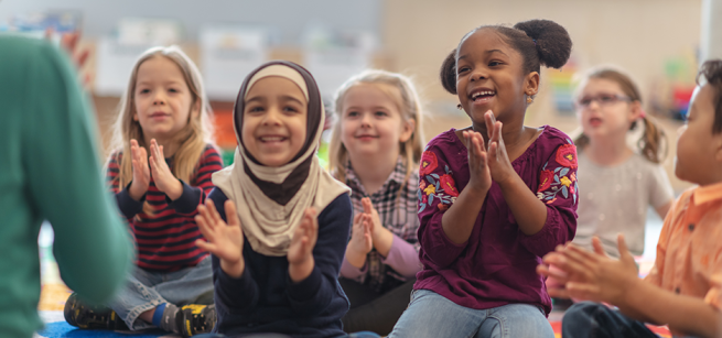 6 small children clapping, smiling, and sitting on the ground facing an adult.