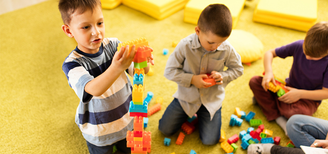 Three young children are playing with colorful building blocks on a soft, yellow carpet.