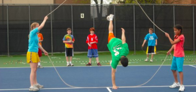 People holding a jump rope on a tennis court, man in center doing a handstand