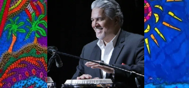 A photo of Louie Cruz Beltran drumming with bright island artwork on either side of him