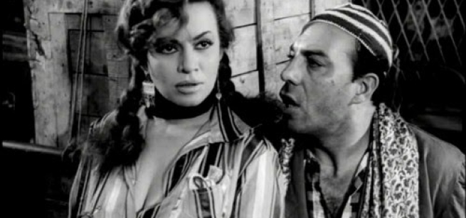 Egyptian man in a skull cap and a woman in pigtails standing next to a fence and wooden box