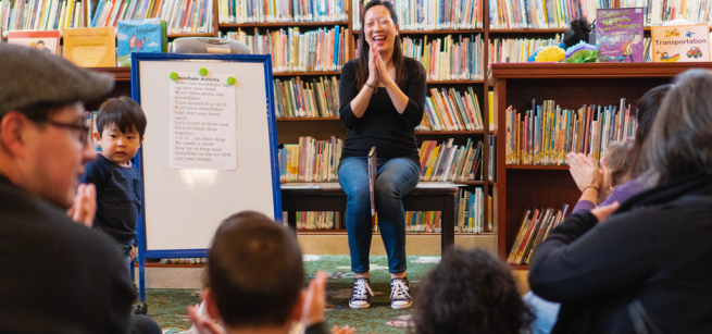 A woman sitsin front of a group of toddlers and adults clapping her hands. A white board is next to her facing the crowd. 