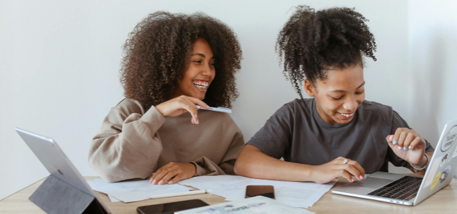 Two teens sitting at a table with laptops smiling and laughing.