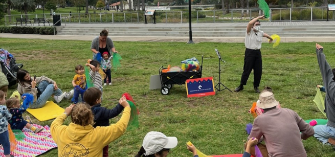 masked librarian leads group of toddlers and caregivers seated on a grassy lawn in singing and waving scarves