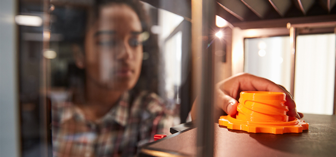 A woman examines a partially printed orange 3D object inside a 3D printer. She appears focused, wearing a plaid shirt, with soft lighting in the background.