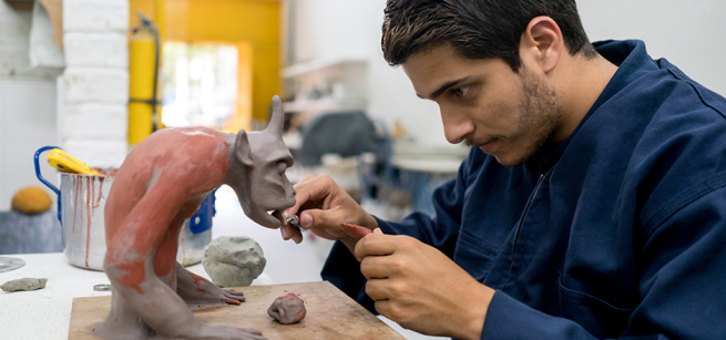 A man sculpts a fanciful creature from clay using pottery tools.