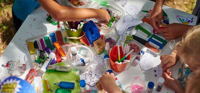 An assortment of craft supplies, paints, markers and hands with scissors cutting paper.
