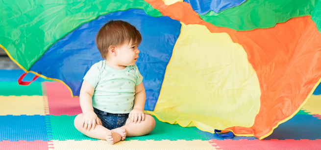 a baby sits on a colorful mat, with a colorful parachute waving behind them