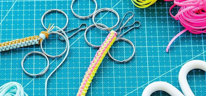 The image shows a boondoggle keychain surrounded by all the craft supplies used to make it such as multi-colored gimp string, scissors, and scattered key rings.