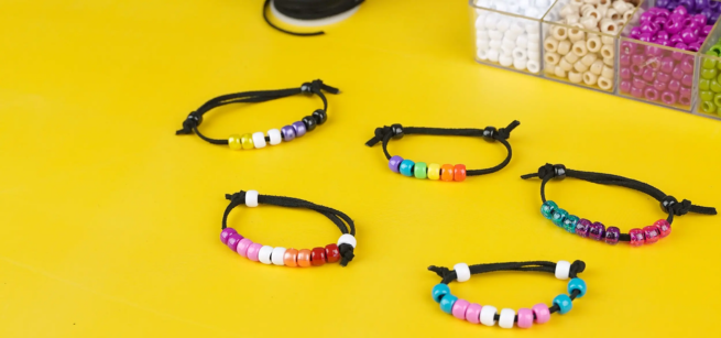 Colorful beaded bracelets on black cords are arranged on a vibrant yellow surface. A box of assorted beads is partially visible in the background.