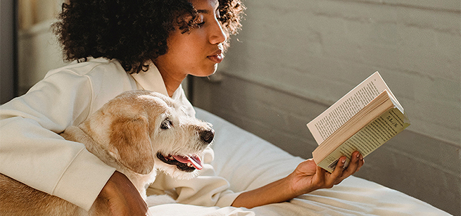 A woman reads a book on a bed with her arm around a dog.