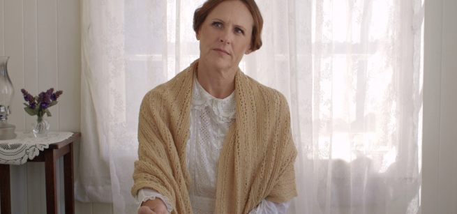 Molly Shannon dressed as Emily Dickinson sits in a sunlight room holding a pen