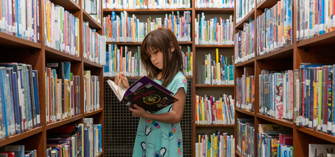 Head & shoulder view of brunette, white girl, reading book her in arms, surrounded by book-filled shelves