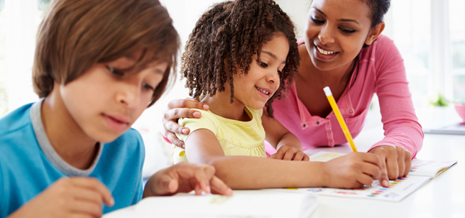 Adult assisting a child with written homework while another student works nearby