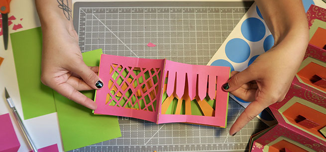 hands displaying pop up book art project, scissors and paper on table