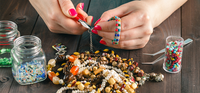 Hands with painted red nails using tools and beads to make a jewelry piece.