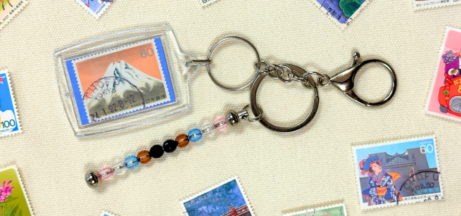 A keychain made of a postage stamp and beads is surrounded by a scattering of postage stamps.