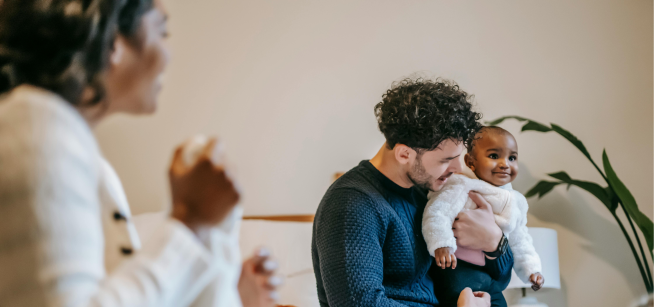 A man is holding a smiling baby while a woman looks on.